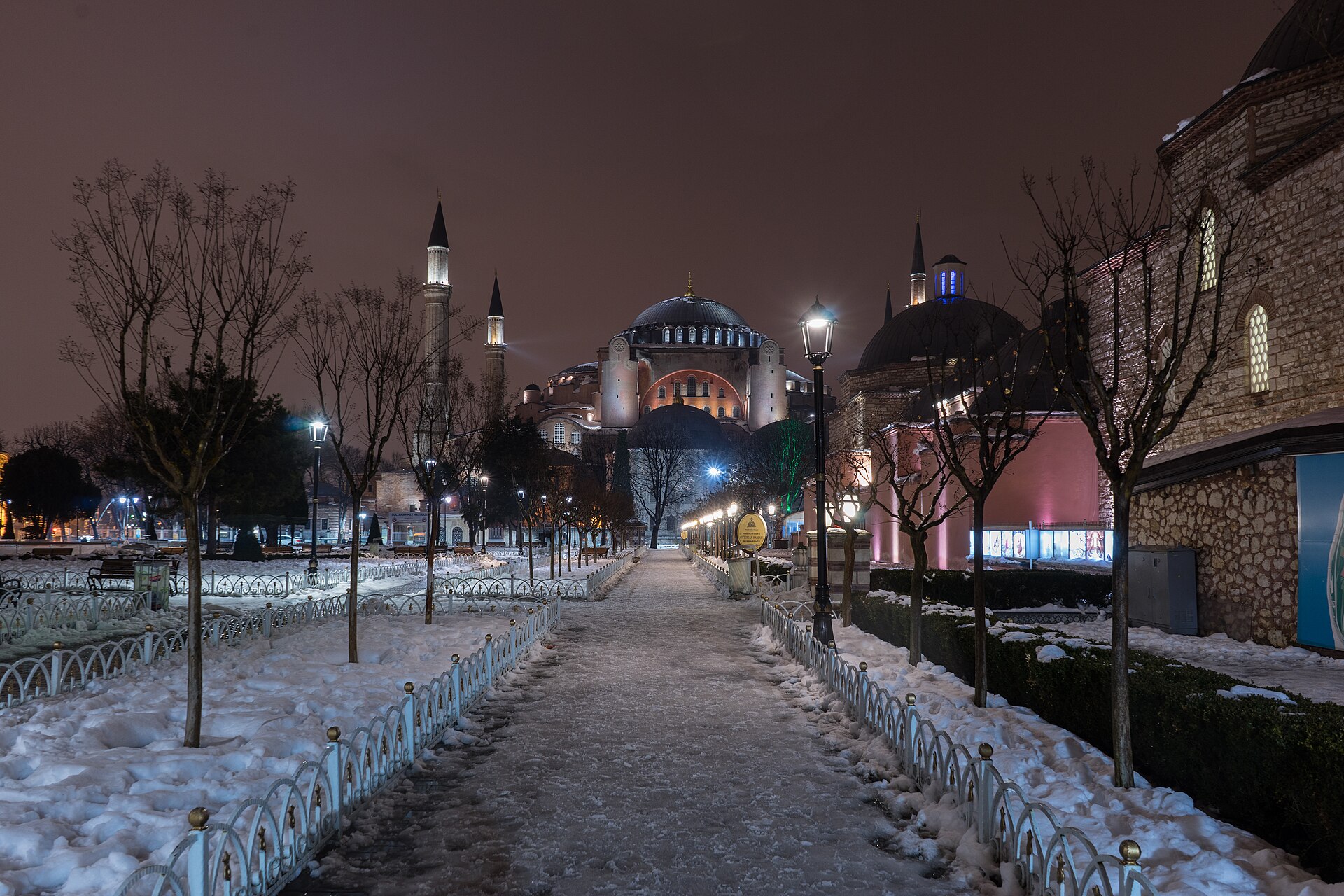 A snowy pavement and trees beside Hagia Sophia in Sultanahmet at night with streetlights