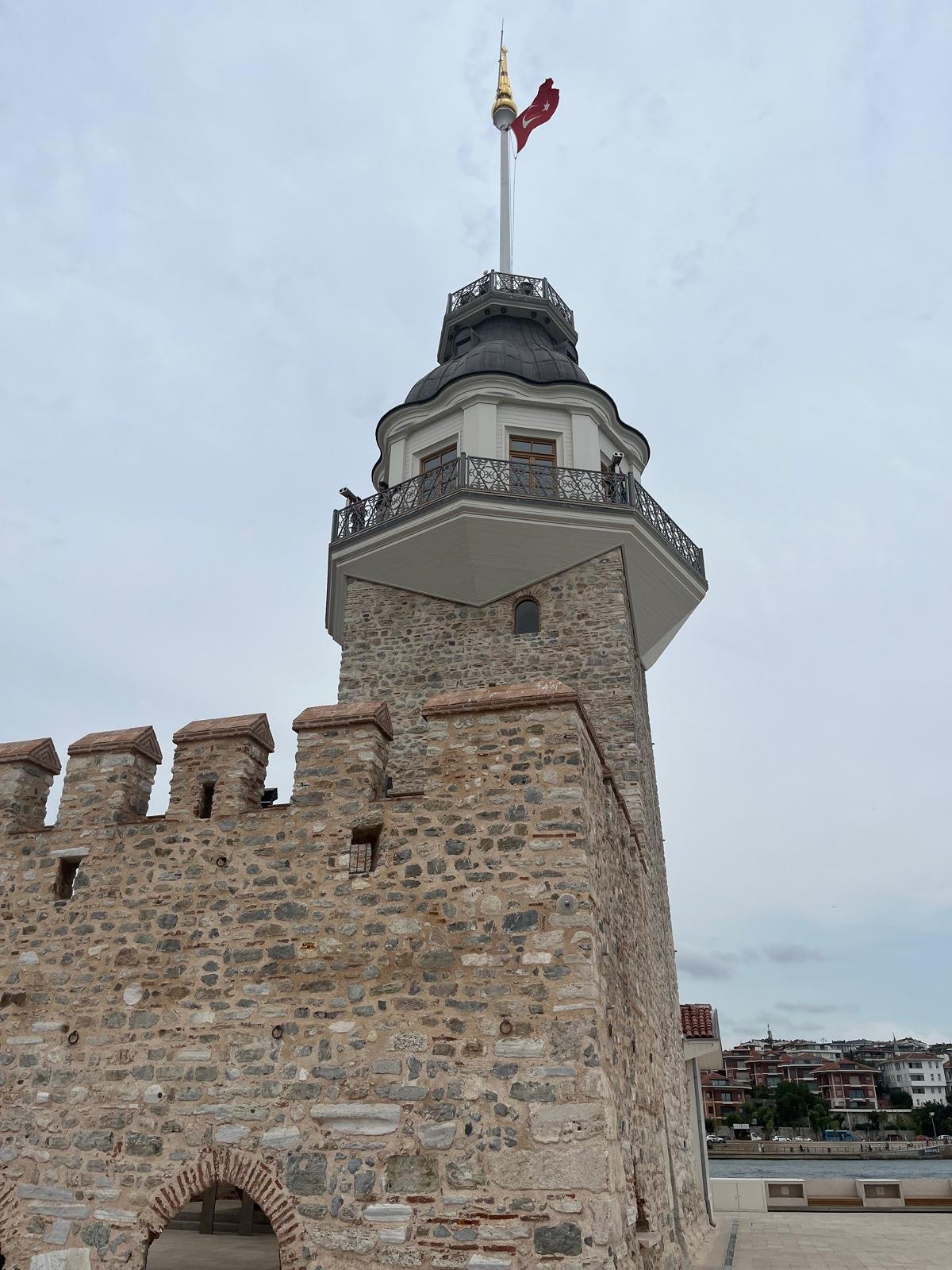 Stone battlements and the tower's balcony and grey dome seen from the island, with a flag on top